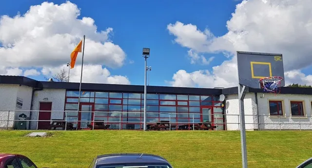 L'edificio moderno della Scoil Ruáin con un canestro da basket si trova su un prato verde sotto un cielo azzurro.