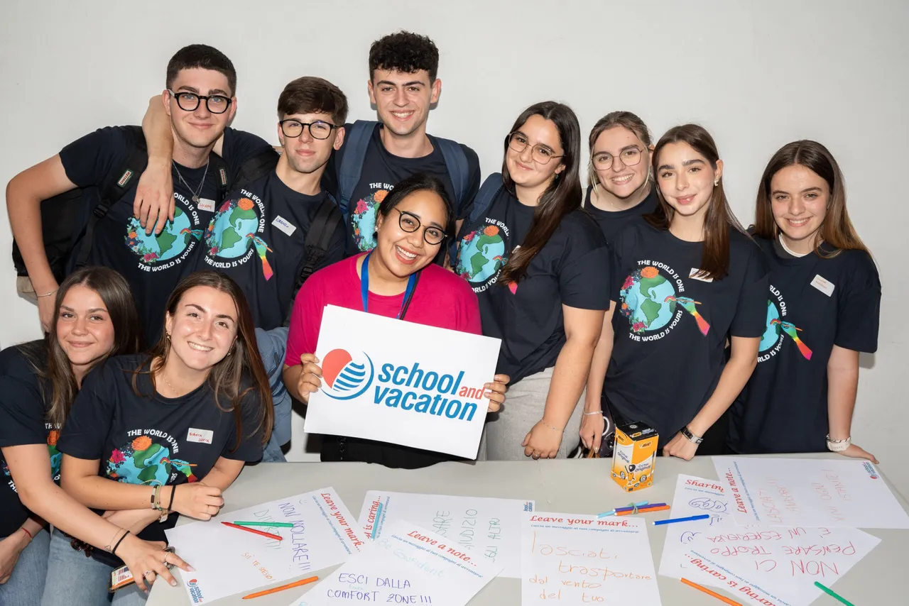 Gruppo di studenti in maglietta sorride con un cartello School and Vacation a un workshop