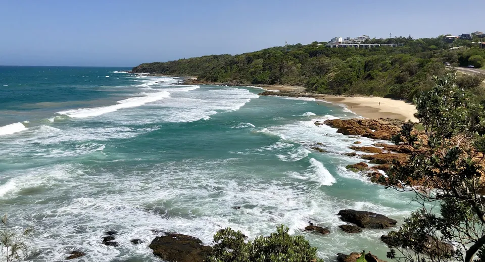 Un paesaggio costiero con spiaggia di sabbia, onde e una scogliera verdeggiante vicino alla Yeppoon State High School.