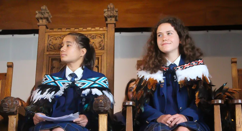 Due studentesse del Wellington East Girls’ College indossano abiti tradizionali davanti a una struttura in legno con intagli.