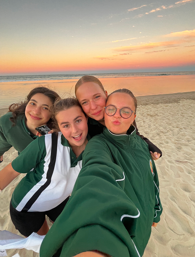 Cuatro estudiantes con uniforme escolar verde se hacen un selfie en la playa al atardecer