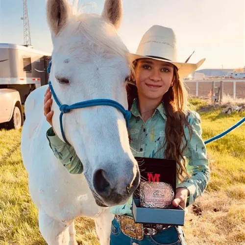 Una mujer con atuendo de cowgirl está junto a un caballo blanco en un prado frente a las montañas en los terrenos de Foremost School.