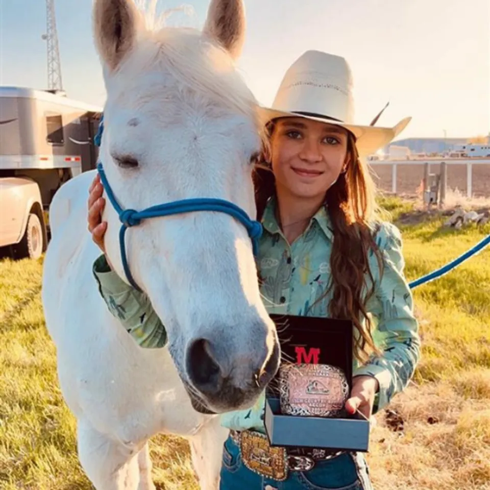 Una donna in outfit da cowgirl è in piedi accanto a un cavallo bianco su un prato davanti alle montagne sul terreno della Foremost School.