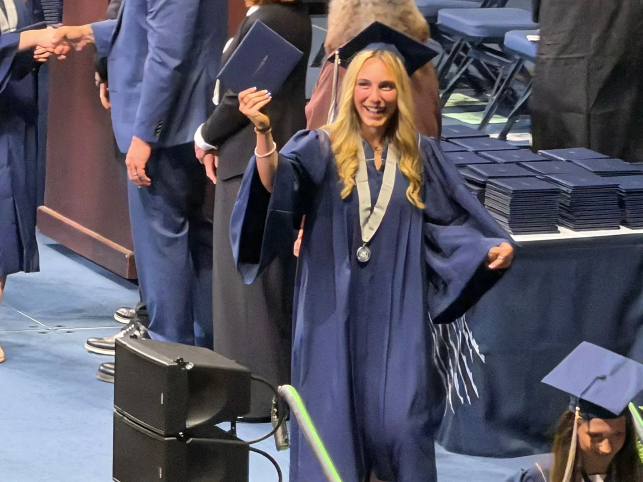Graduada con toga y birrete azules levantando su diploma en la ceremonia de graduación