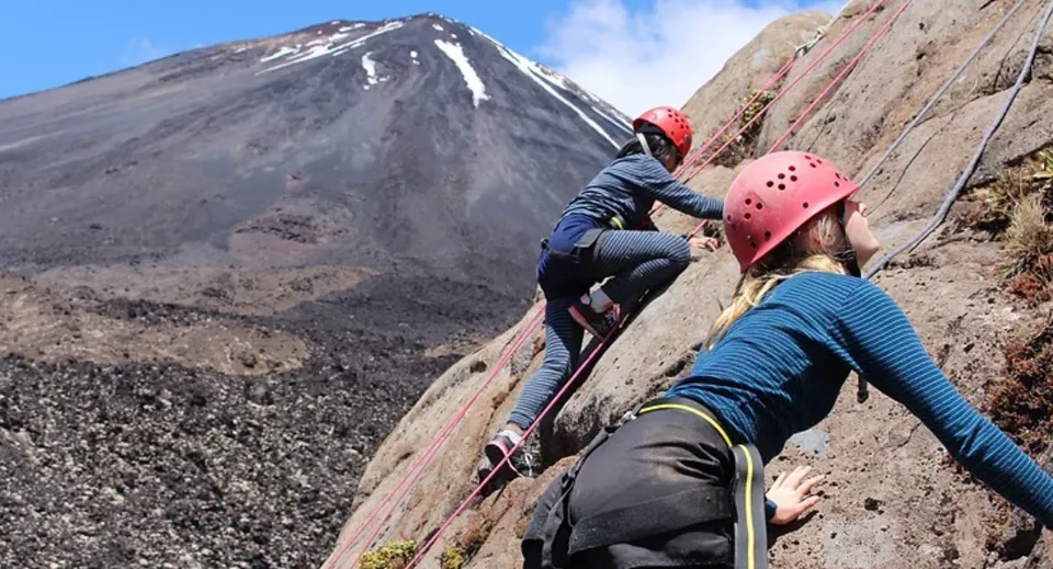 Due studenti della Freyberg High School si arrampicano con l'attrezzatura su una parete rocciosa con una montagna innevata sullo sfondo.