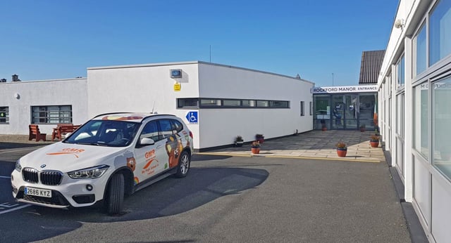Una BMW X1 bianca è parcheggiata davanti a un grande edificio bianco nel campus della Rockford Manor School sotto un cielo limpido.