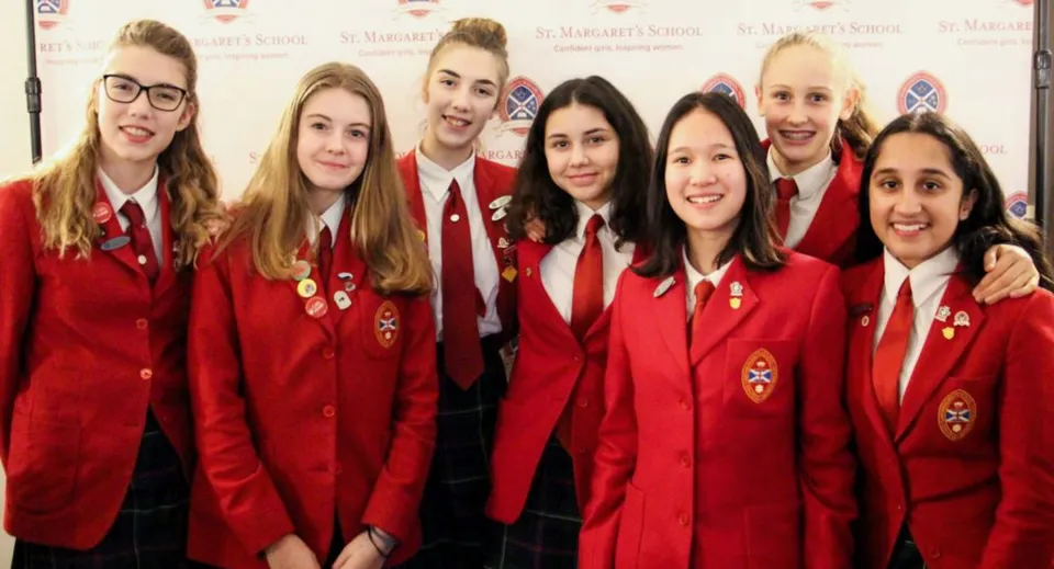 Un gruppo di studentesse e studenti della St. Margaret’s School posa in uniformi rosse davanti a uno sfondo con il logo.
