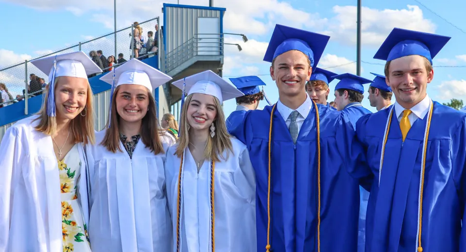 Un gruppo di diplomati della Sir Guy Carleton Secondary School in toga blu sta davanti a un prefabbricato sotto un cielo nuvoloso.