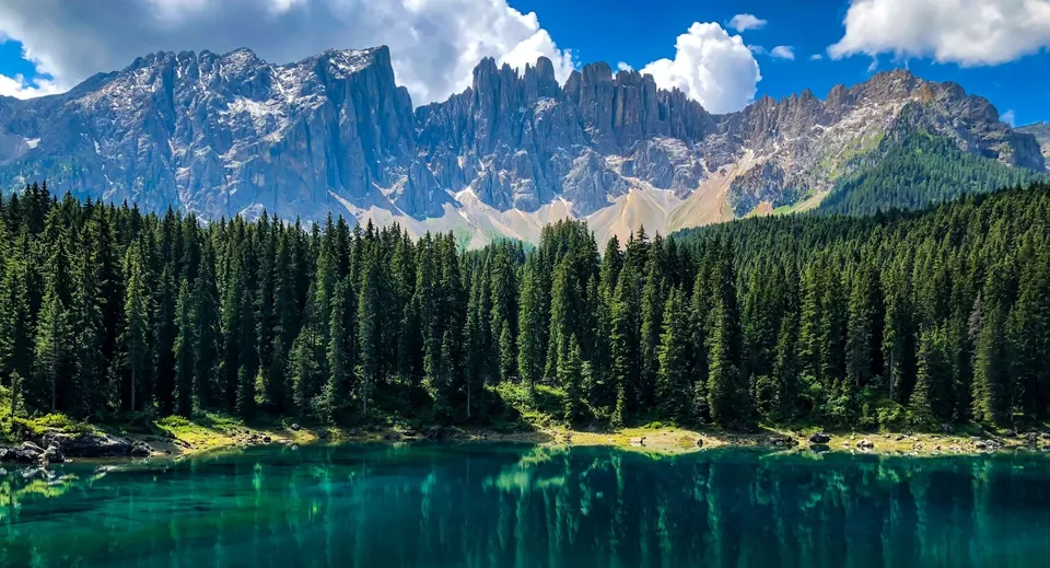 Un lago di montagna turchese con cime innevate e una foresta di sempreverdi in primo piano.