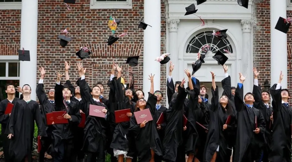 I diplomati della Princeton International School of Mathematics & Science lanciano in aria i loro tocchi da diploma davanti a un edificio in mattoni.