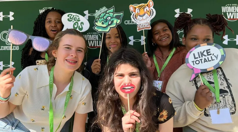 Un gruppo di studentesse sorridenti posa davanti a un pannello fotografico con grafiche nel campus della Oldfields School.