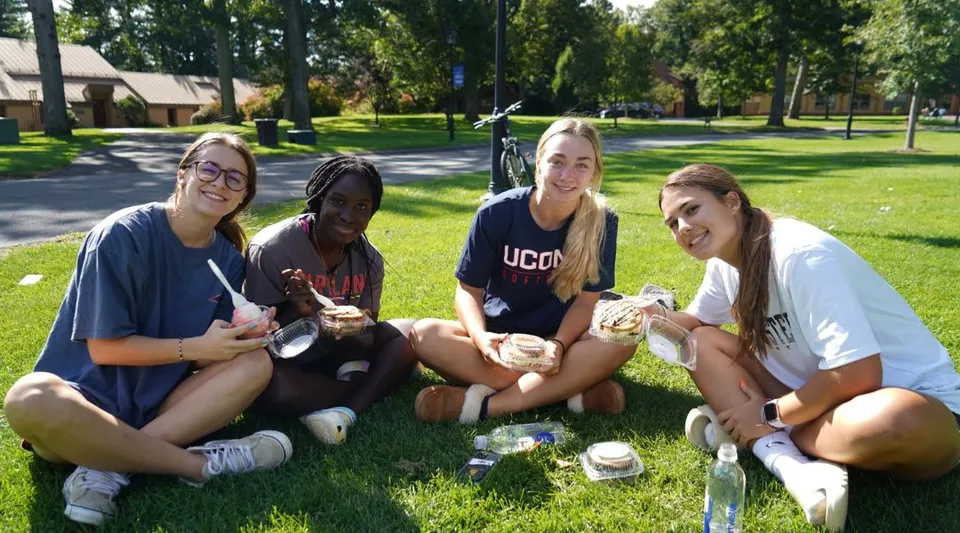 Quattro studentesse sono sedute sull'erba nel parco vicino alla Westminster School con alberi ed edifici sullo sfondo.