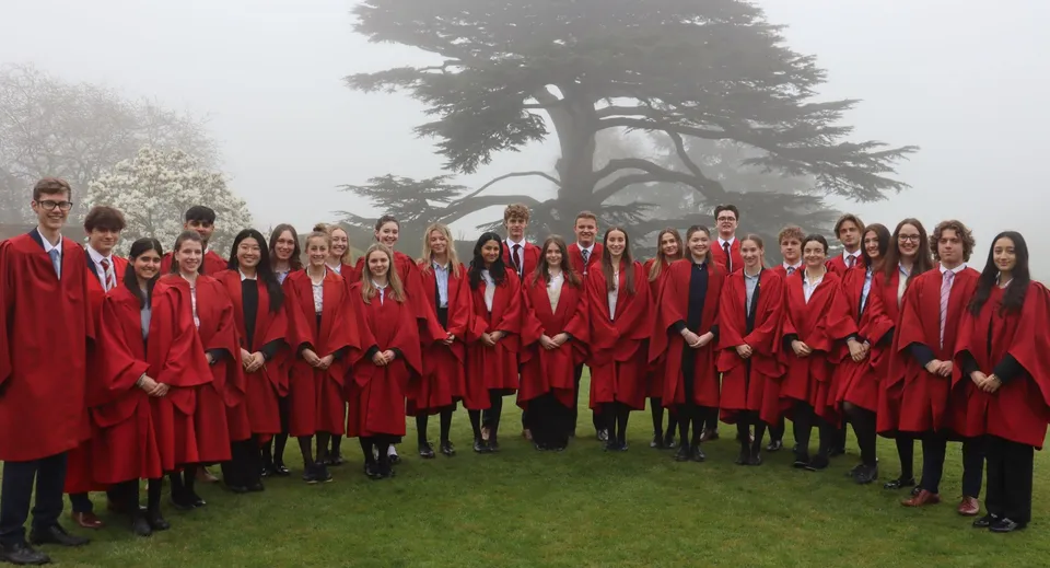 Un grande gruppo di studenti della Westonbirt School con tuniche rosse si trova su un campo erboso davanti a un grande albero nella nebbia.