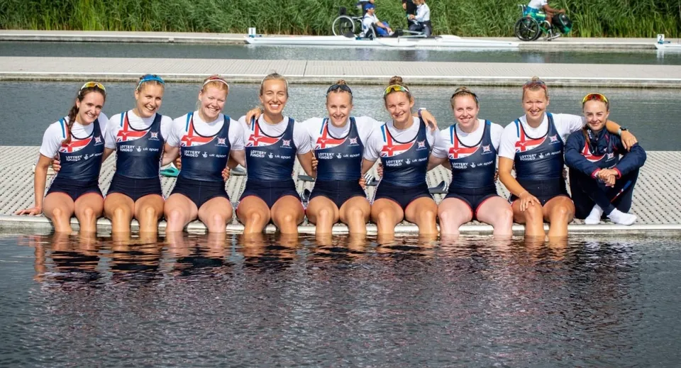 Un gruppo di atlete della Durham School, vestite con la stessa divisa, siede in acqua davanti a un'area erbosa con ciclisti sullo sfondo.