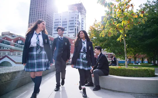 Un grupo de estudiantes de la City Vancouver Academy High School camina junto por el campus escolar con edificios altos y árboles.