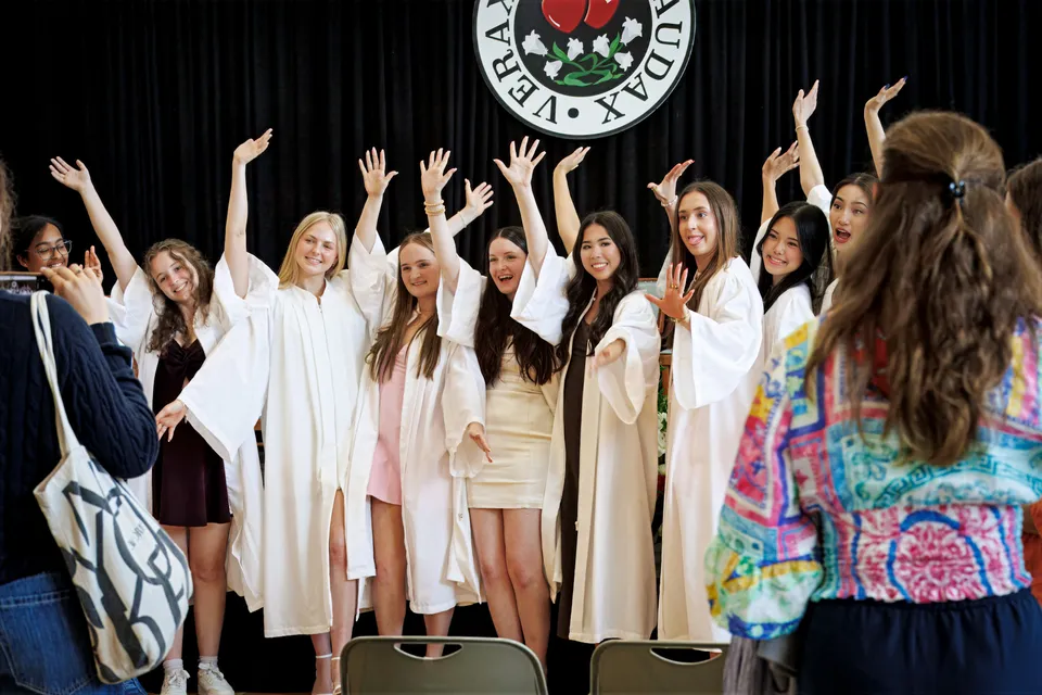 Un gruppo di giovani studentesse della Sacred Heart School of Halifax si trova su un palco in tuniche bianche con le braccia sollevate davanti a un grande logo.