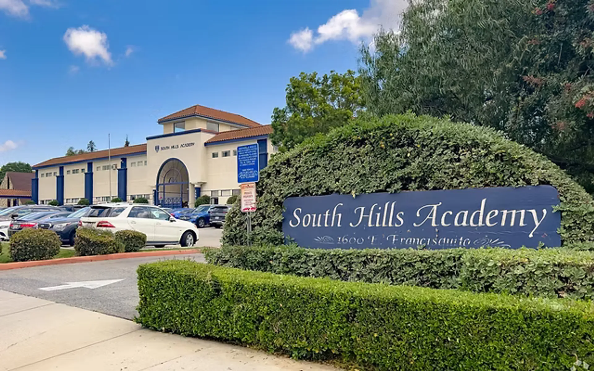 El edificio de la South Hills Academy detrás de un seto, con el cartel con el nombre del colegio y el aparcamiento en primer plano.