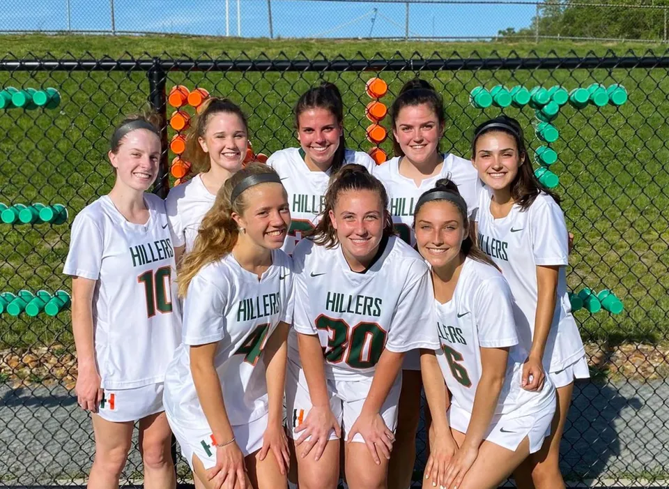 Un gruppo di giovani atlete in uniformi bianche in posa davanti a una recinzione verde nel campus della Hopkinton High School.