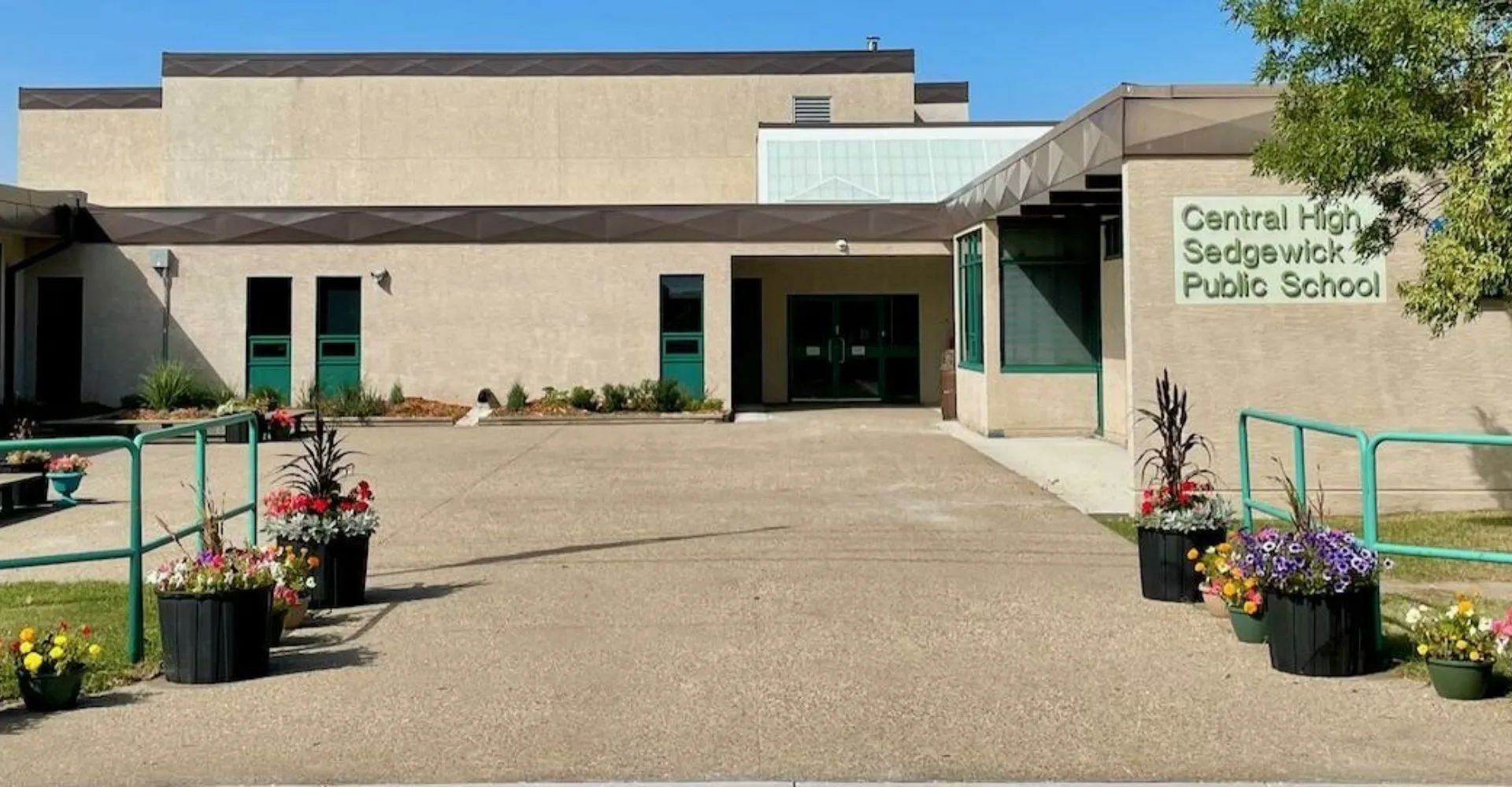 L'edificio scolastico moderno della Central High Sedgewick Public School con ingresso pavimentato e fiori è circondato da alberi.