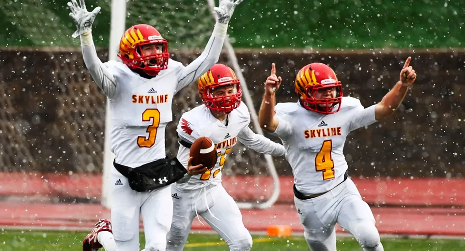 Tre giocatori di football americano in divisa bianca e caschi rossi festeggiano sul campo bagnato delle St. Vrain Valley Schools.