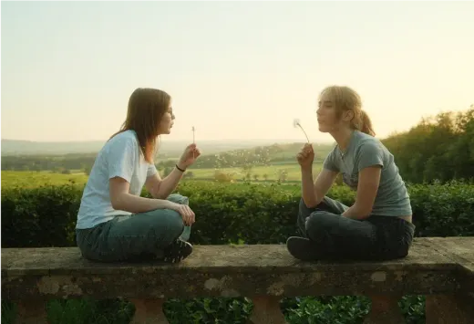 Zwei Mädchen sitzen bei Sonnenuntergang auf einer Mauer und pusten Pusteblumen in der englischen Landschaft