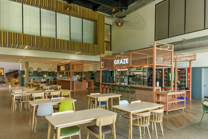 Comedor del campus con mesas de madera, sillas claras y puestos de comida al fondo