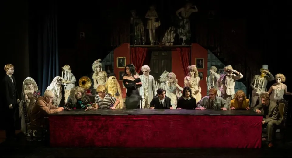 Un gruppo di persone in costume si trova sul palco della Ipswich School davanti a un sipario rosso con uno sfondo decorativo.