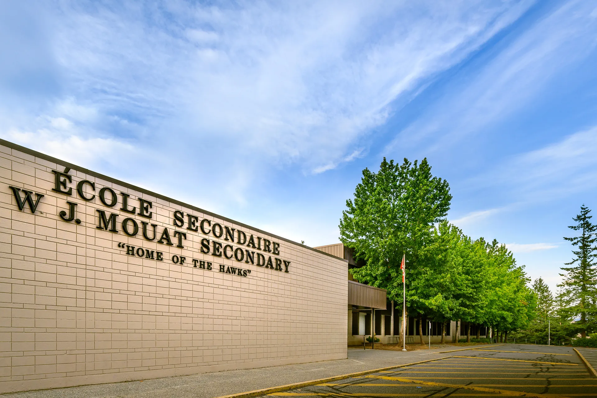 Il grande edificio della W.J. Mouat Secondary School è circondato da alberi verdi e si trova sotto un cielo blu con nuvole.
