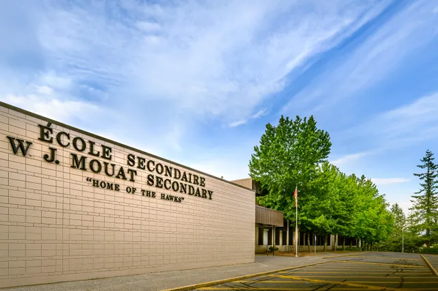 Il grande edificio della W.J. Mouat Secondary School è circondato da alberi verdi e si trova sotto un cielo blu con nuvole.