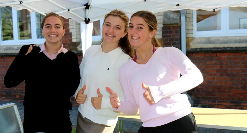 Tre studentesse sorridenti sono insieme davanti a un edificio con vetrate nel campus della Queen Anne’s School.