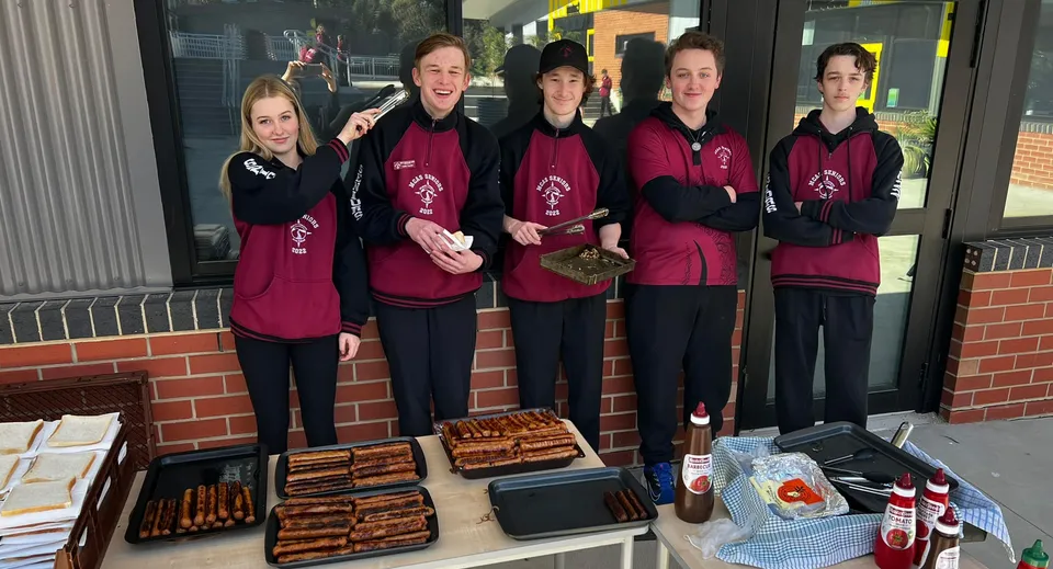 Cinque studenti e studentesse della Mount Compass Area School sono in piedi in divise coordinate color bordeaux davanti a un negozio di alimentari e attrezzature.