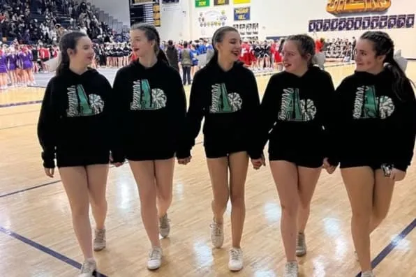 Cinco chicas jóvenes caminando cogidas de la mano con sudaderas negras en un gimnasio escolar