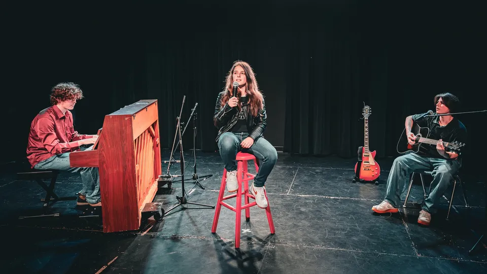 Tre studenti della Pioneer School Port Coquitlam si esibiscono su un palco con pianoforte, chitarra e canto.