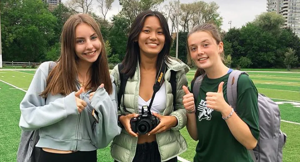 Tre studentesse sorridono immerse nel verde del campus dell'Etobicoke Collegiate Institute e mostrano il pollice all'insù.