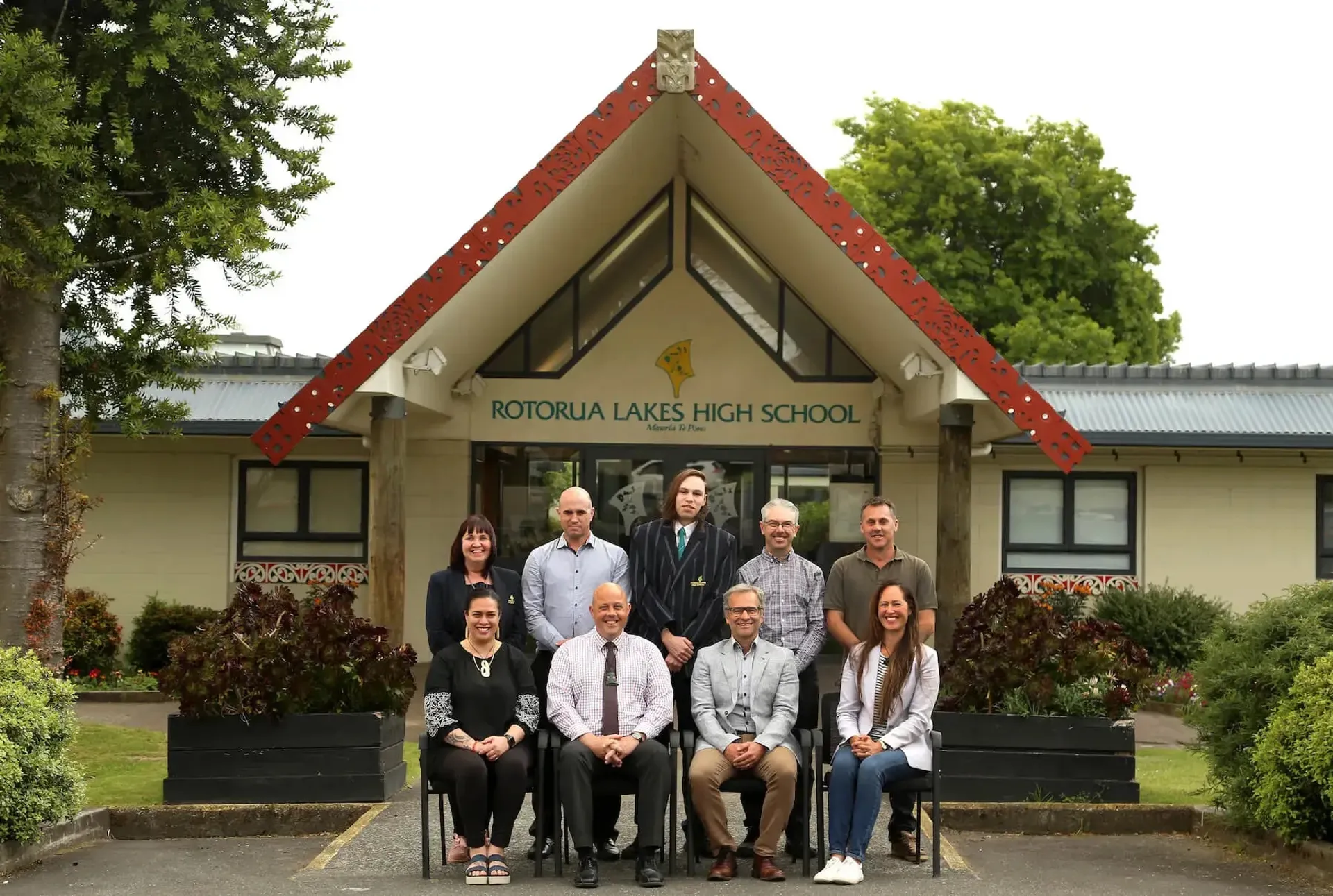 Membri del personale della Rotorua Lakes High School davanti all'edificio scolastico con un caratteristico tetto triangolare, circondato dal verde.