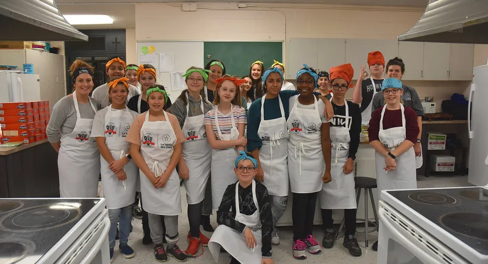 Studenti della Heritage Regional High School indossano grembiuli bianchi e lavorano in una cucina scolastica con attrezzature da cucina.
