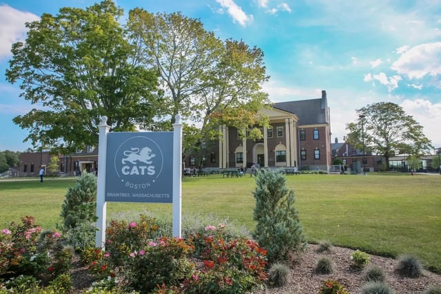 Ein Schild der CATS Academy Boston steht auf einer Grünfläche vor dem großen Backsteingebäude der Schule.