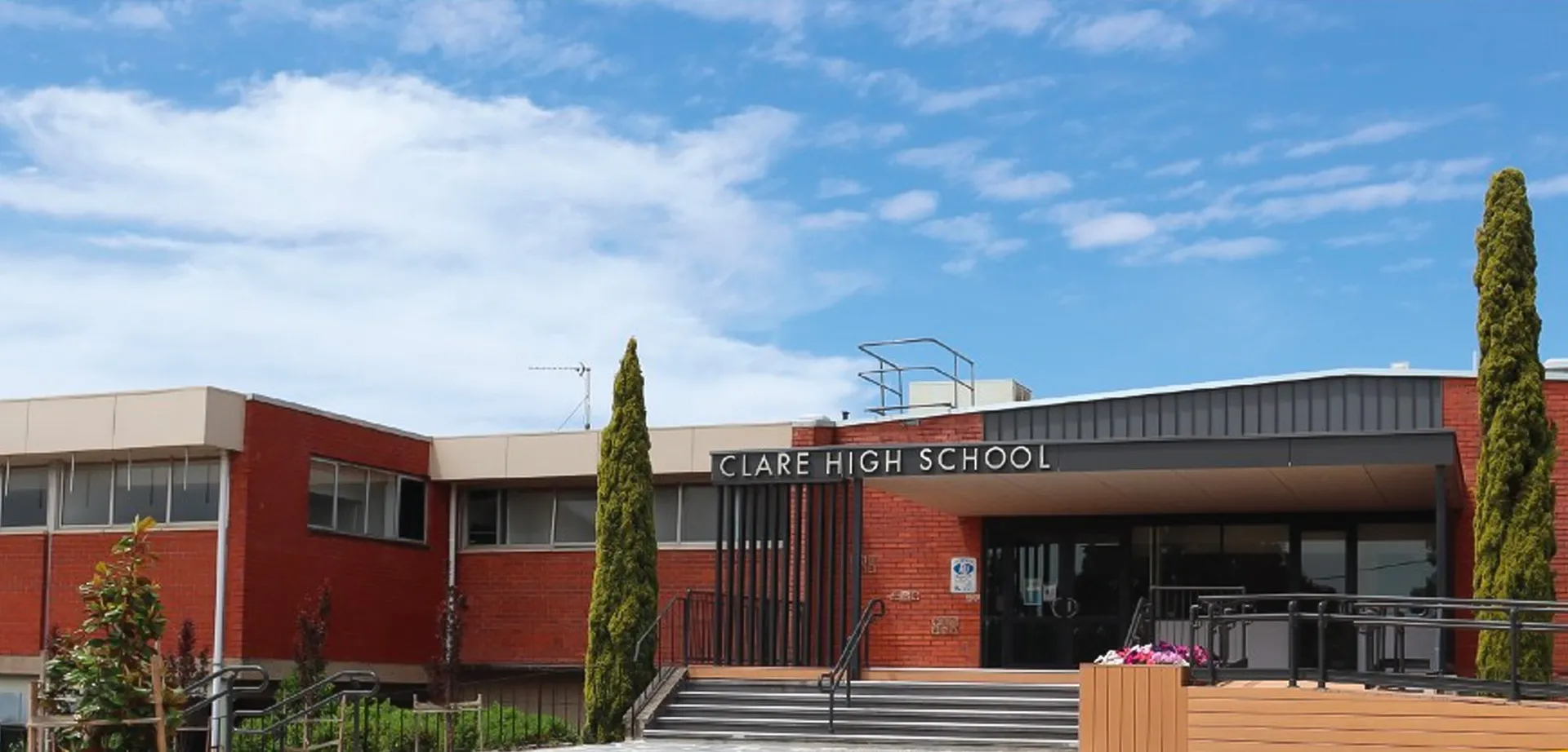 L'edificio in mattoni rossi della Clare High School è circondato da alberi e si trova sotto un cielo blu nuvoloso.