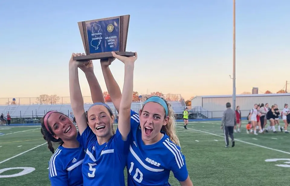 Tre studentesse della Shore Regional High School festeggiano con un trofeo sul campo sportivo davanti allo skyline della città.