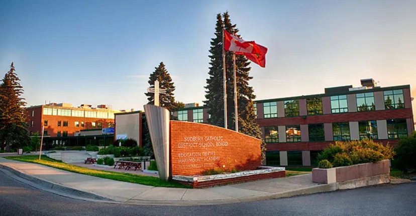 Una grande bandiera rossa si trova davanti a un monumento nel campus della Marymount Academy, con edifici moderni e alberi sullo sfondo.