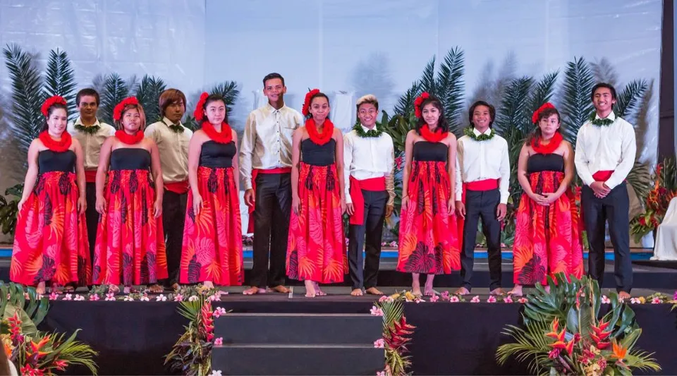 Studenti della Hawaiian Mission Academy sul palco con costumi rossi e bianchi su uno sfondo tropicale.