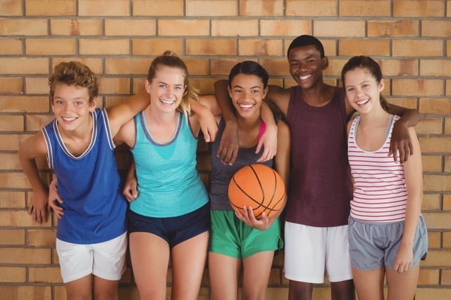 Fünf Jugendliche in Sportkleidung mit Basketball stehen Arm in Arm vor einer Backsteinwand