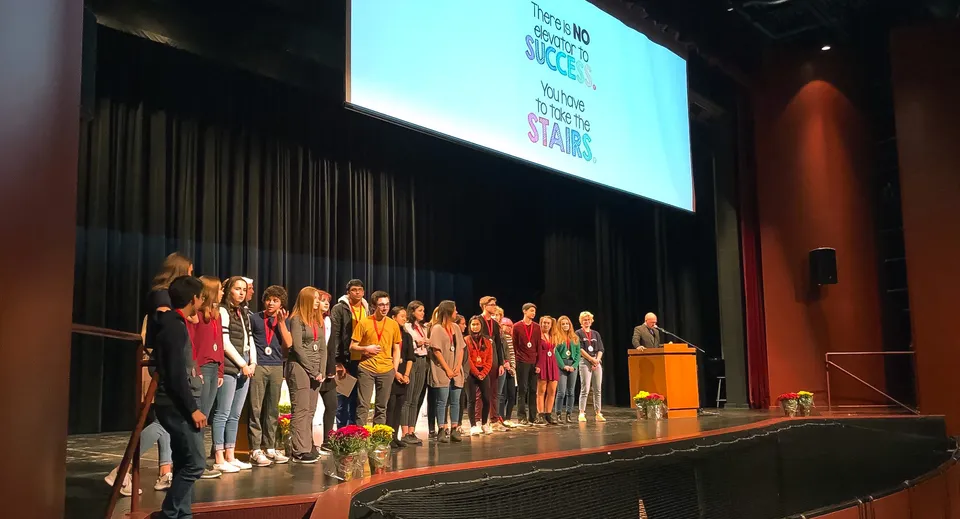 Studenti della Sacred Heart High School sono sul palco davanti a un grande schermo di proiezione con del testo.