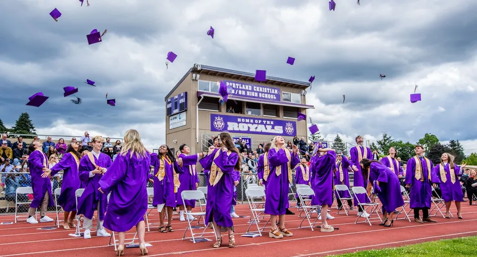 Diplomati della Portland Christian High School festeggiano con toghe viola e cappelli lanciati in aria sul campo sportivo.