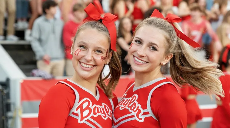Due cheerleader sorridenti in divisa rossa nel campus della Baylor School davanti a una folla di persone.