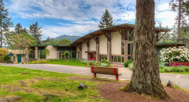Una casa in legno in stile lodge si trova nel campus della St. Margaret’s School, circondata da alti pini e montagne sullo sfondo.