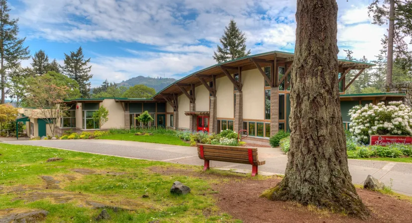 Una casa in legno in stile lodge si trova nel campus della St. Margaret’s School, circondata da alti pini e montagne sullo sfondo.