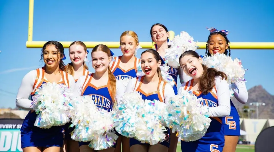 Cheerleader del Phoenix Union High School District in piedi con pompon colorati davanti a un cielo azzurro e un palo della porta giallo.