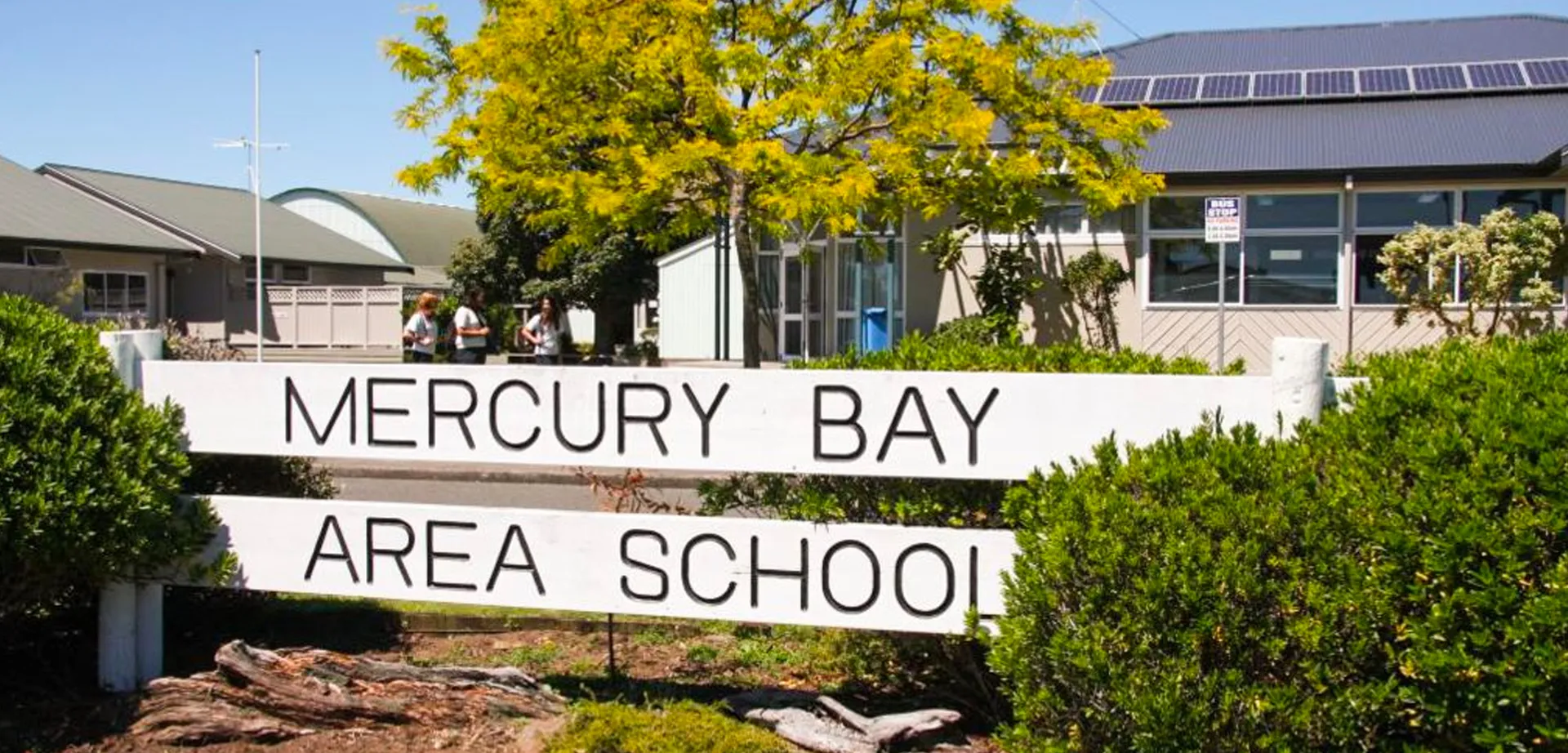 L'insegna della Mercury Bay Area School si trova davanti agli alberi con un edificio scolastico sullo sfondo.