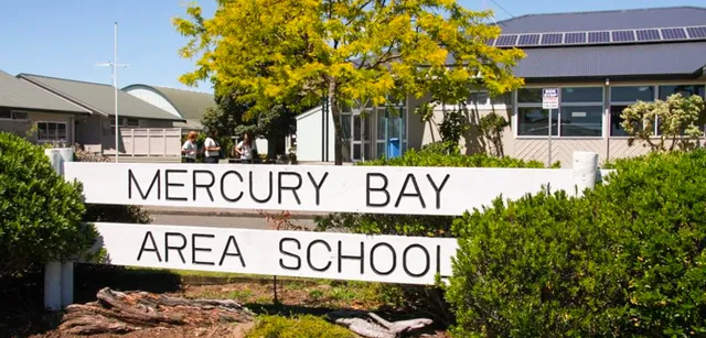 L'insegna della Mercury Bay Area School si trova davanti agli alberi con un edificio scolastico sullo sfondo.
